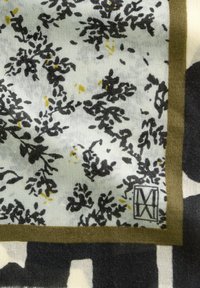 Sjaal met bloemenpatroon in zachte stof. Bevat zwarte en gele motieven op een lichte achtergrond, omzoomd met een olijfgroene rand. Label zichtbaar.