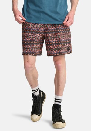 Mann trägt schwarz-orange gemusterte Shorts, blaues T-Shirt, weiße Socken mit schwarzen Streifen und schwarze High-Top-Sneaker, steht vor weißem Hintergrund.