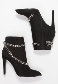 Bottines en daim noir avec talon aiguille, fermeture éclair et accents de chaîne argentée enroulés autour de la cheville.