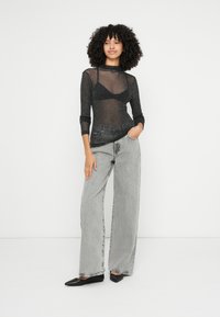 Top nero trasparente a maniche lunghe indossato sopra un reggiseno nero, abbinato a jeans grigio chiaro a gamba larga e ballerine nere su uno sfondo semplice.