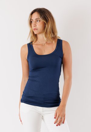 Top aderente blu navy con scollatura tonda, realizzato in materiale morbido, presenta un design laterale leggermente arricciato. Pantaloni bianchi e gioielli semplici.