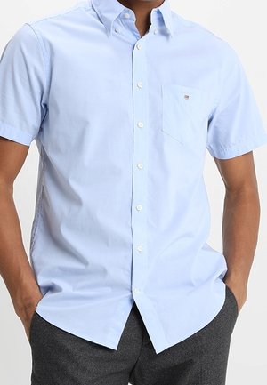 Mann trägt ein hellblaues Kurzarm-Hemd mit Knöpfen und Brusttasche sowie dunkelgraue Hose, Hände in den Taschen.