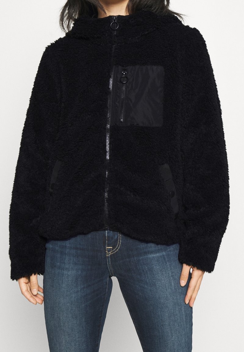 Svart fuskpäls zip-jacka med en framficka med dragkedja, huva och sidoknappar. Texturerad och varm för svalare väder.