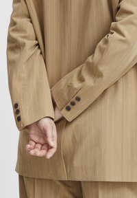 Beige csíkos öltönyzakó gombos mandzsettával; sima szövettextúra; kéz a hát mögött, diszkréten bemutatva és laza tartással.