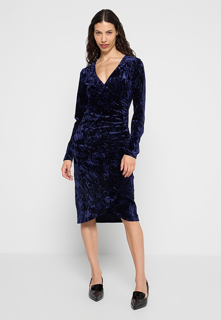 Robe en velours bleu marine avec un décolleté en V, des manches longues et une silhouette ajustée. Présente un design enveloppé et une surface texturée.
