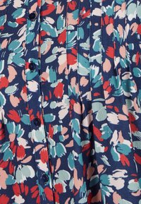 El tejido del vestido floral presenta patrones abstractos en rojo, azul verdoso y rosa sobre un fondo marino; incluye botones y detalles con pliegues.