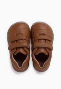 Scarpe in pelle marrone con forma arrotondata, dotate di due cinturini in Velcro e una suola texture. L'interno presenta un motivo circolare rosso.
