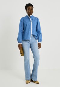 Camisa de mezclilla azul de manga larga con encaje a lo largo del frente y las mangas, combinada con vaqueros acampanados azul claro y botines con tacón color cuero claro.