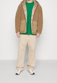 Chaqueta marrón de sherpa con forro de satén, que presenta grandes bolsillos. Camiseta verde debajo, combinada con pantalones de pana beige y zapatillas multicolores.