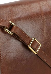 Dettaglio di una borsa a tracolla in pelle marrone con superficie texturizzata, tracolla regolabile con fibbia in metallo dorato e dettagli cuciti.