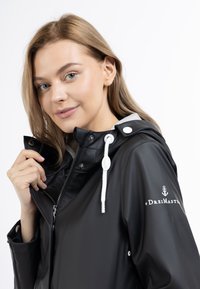 Giacca impermeabile nera con cappuccio grigio, cordini bianchi e zip frontale. Presenta un logo e snap sulle maniche per la regolabilità.