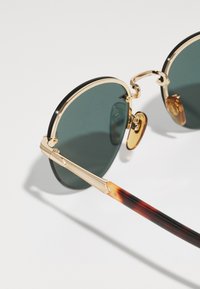 Ochelari de soare rotunzi cu lentile de culoare verde închis, ramă din metal aurit și brațe cu model țestoasă. Prezintă accente decorative pe tâmple.