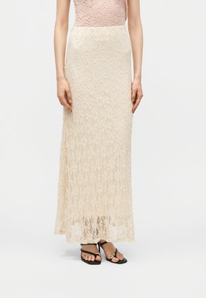 Femme portant une longue jupe en dentelle beige associée à des sandales noires à brides avec un détail chaîne dorée, debout devant un fond uni.