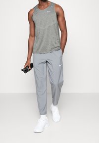 Szary podkoszulek Nike o lekkiej, oddychającej teksturze; w połączeniu z szarymi spodniami dresowymi z białym logo. Trzyma czarną butelkę na wodę.