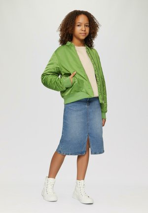 Groene satijnen bomberjack met geribde manchetten, beige top, spijkerrok met een split aan de voorkant, en witte hoge sneakers. Eenvoudige en casual uitstraling.