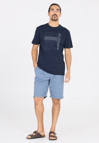 Merin t-paita, jossa on painettu teksti, yhdistettynä vaaleansinisiin, käännettyihin shortseihin ja ruskeisiin sandaaleihin. Rentouttava asu, jossa on mukava istuvuus ja standardimateriaalit.