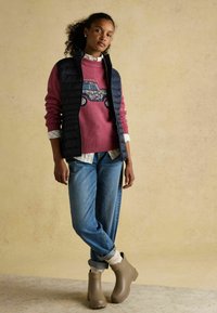 Rozā džemperis ar ziedu kravas automobiļa dizainu, tumši zilā iešūto vesti, gaiši zilos džinsos, krēmveida zeķēs un brūnos potīšu zābakos uz teksturēta bēša fona.