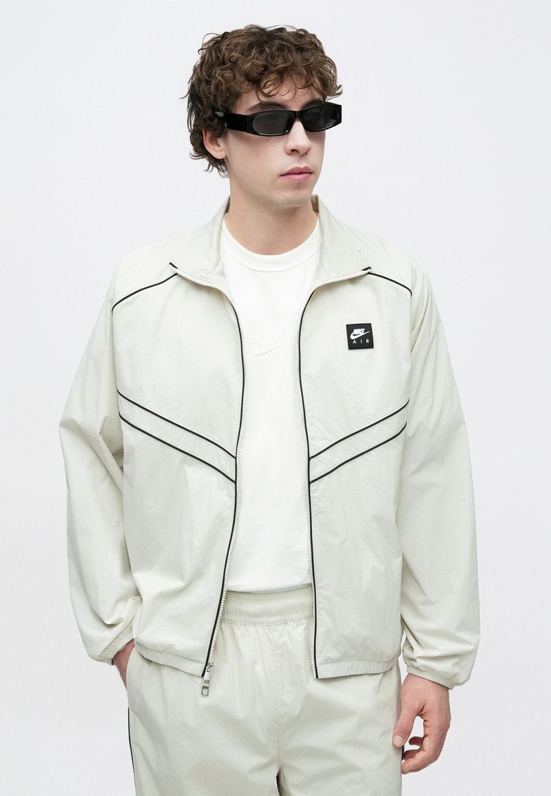 Chaqueta deportiva gris claro con detalles en negro y logo de Nike, combinada con una camiseta blanca. El modelo lleva gafas de sol rectangulares oscuras.
