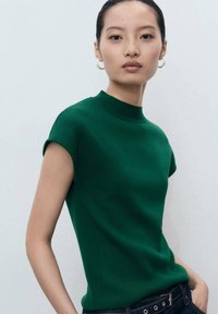 Green short-sleeve top with a high neckline, made of ribbed fabric. Features a fitted shape and worn with dark denim, showcasing a minimalist style.