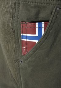 Olivgrüner Stoff mit einem Patch, der eine norwegische Flagge in Rot, Weiß und Blau zeigt, sichtbar in einer Seitentasche mit Messingbeschlägen.