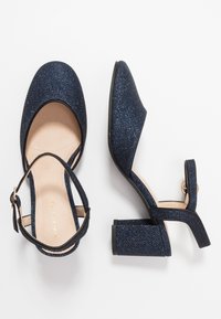 Mörkblå glittrande klackar med rund tå, ankelrem och blockklack. Slät inredning med synlig söm längs kanterna.
