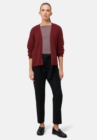 Czerwony miękki kardigan noszony na paskowanej koszuli, zestawiony z czarnymi, dopasowanymi spodniami i czarnymi loafersami, o luźnym kroju i otwartym przodzie.