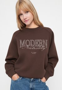 Sweat-shirt marron avec le texte "MODERN Neutrality" en blanc, texture douce, manches raglan et coupe décontractée, associé à un jean bleu.