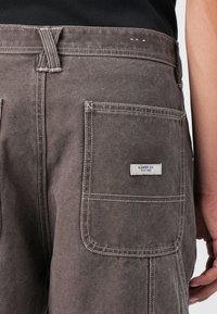 Braune Cargo-Shorts mit einer Gesäßtasche mit Label, verstärkten Nähten und einer strukturierten Stoffoberfläche. Subtile Gebrauchsspuren sind sichtbar.