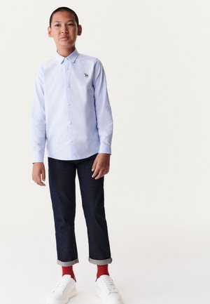 Helles blaues Langarmhemd mit Knopfleiste, dunkle Jeans mit aufgerollten Säumen, rote Socken und weiße Turnschuhe. Das Hemd hat ein kleines Logo.