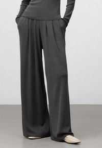 Pantaloni a gamba larga in tessuto grigio scuro, con pinces, due tasche laterali e una texture morbida, abbinati a scarpe beige a punta.