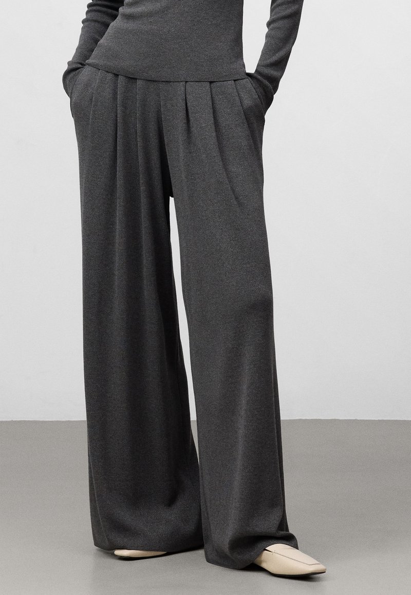 Pantaloni a gamba larga in tessuto grigio scuro, con pinces, due tasche laterali e una texture morbida, abbinati a scarpe beige a punta.
