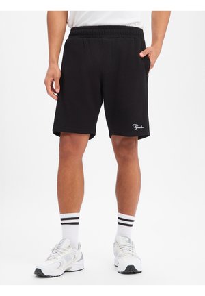 Man draagt zwarte geribbelde shorts met elastische taille en wit logo, witte sokken met zwarte strepen en witte sportschoenen, staat met ontspannen handen.