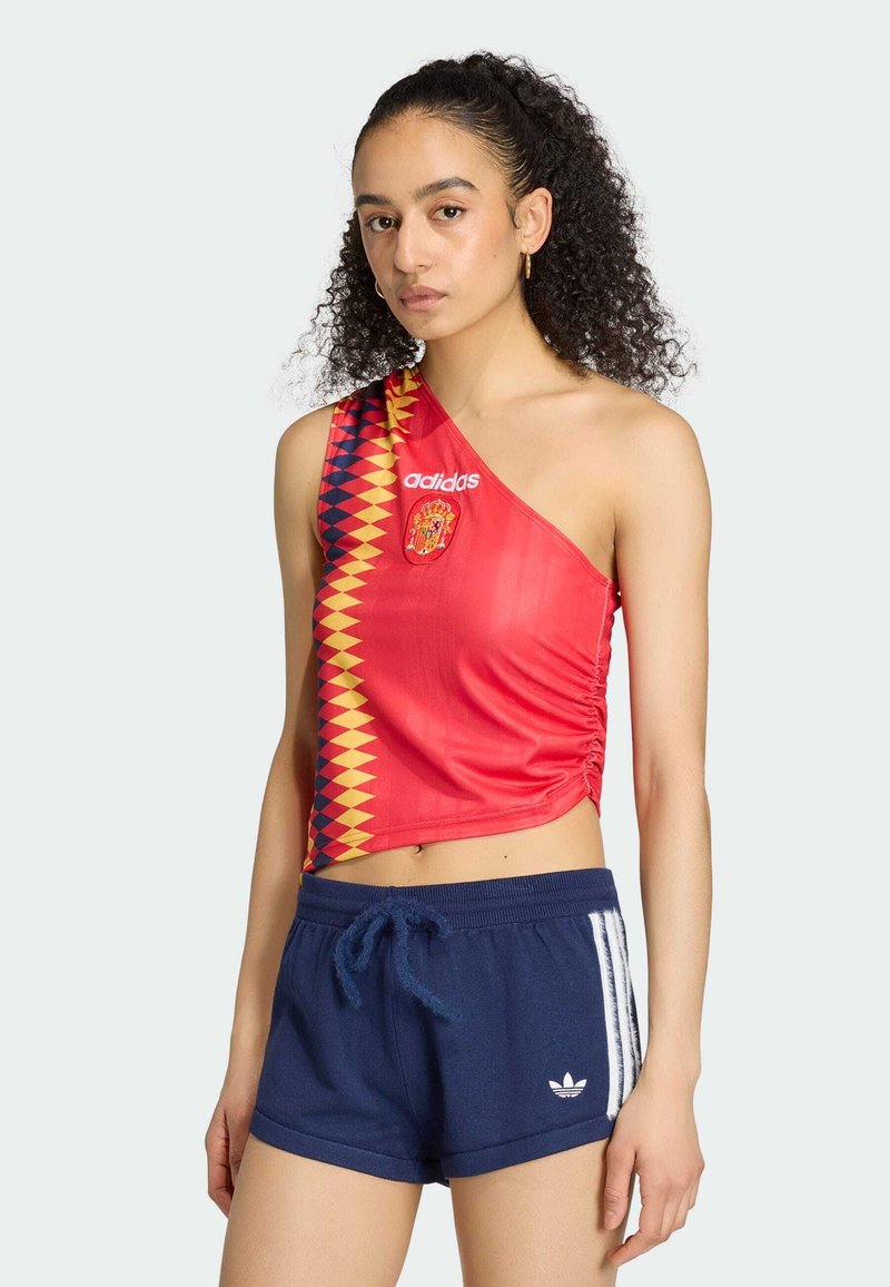 Γυναίκα με σγουρά μαλλιά φοράει κόκκινο τοπ Adidas με έναν ώμο και έμβλημα της Ισπανίας, καθώς και μπλε σορτς Adidas με λευκές ρίγες.
