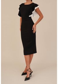 Svart åtsittande klänning med vita volangärmar, knälång, gjord av stretchigt material. Kombinerad med svarta spetsiga klackskor med ankelremmar.