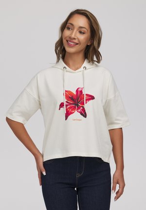 Kobieta w białej bluzie z krótkim rękawem z dużym czerwonym nadrukiem kwiatowym i ciemnoniebieskich dżinsach, uśmiechająca się i pozująca na jednolitym tle.