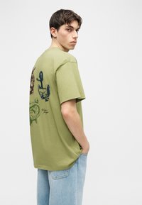 Νέος άνδρας με κοντά σκούρα μαλλιά, που φοράει ανοιχτό πράσινο μπλουζάκι με τύπωμα και ανοιχτόχρωμο μπλε τζιν, κοιτάζοντας προς τα πίσω πάνω από τον ώμο του, με φόντο έναν απλό τοίχο.