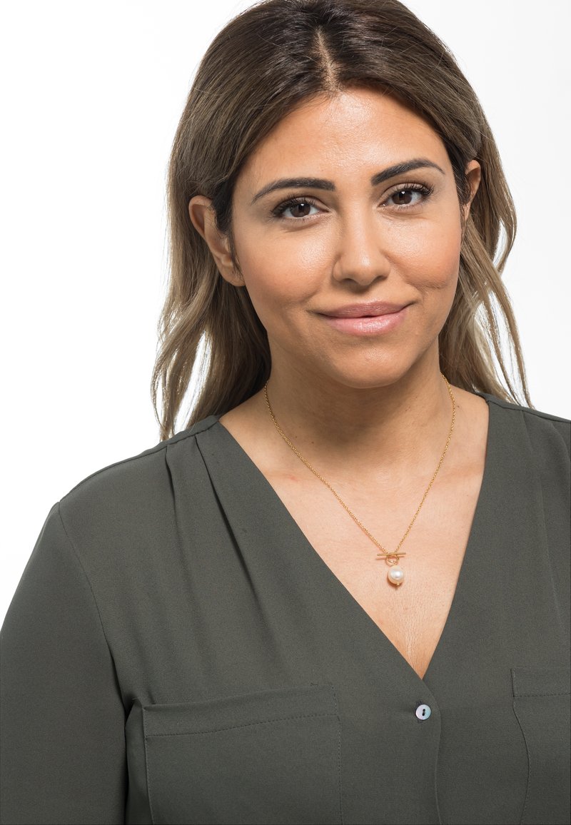 Gouden halsketting met een parelpendant; het model draagt een donkergroene blouse met een V-hals en een borstzakje. Soepele stoftextuur.