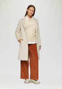 Beige Wollmantel mit Revers, über einem gestrickten hellbraunen Pullover und einem weißen Hemd getragen, kombiniert mit rostfarbenen Cordhosen und beigen klobigen Stiefeln.