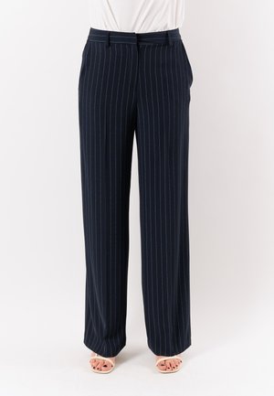 Donna con pantaloni a gamba larga blu navy con sottili righe bianche e tacchi aperti beige su uno sfondo neutro.
