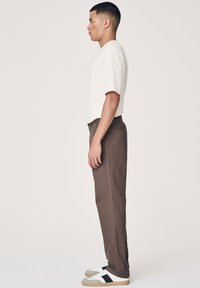 Beige t-shirt med korte ærmer, brune lige bukser og sneakers med grå, sort og hvide detaljer, vist fra en sidevinkel.