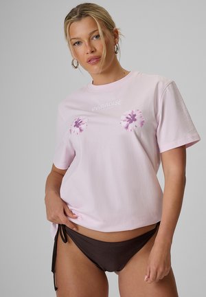 Kobieta w jasnoróżowej koszulce z kwiatowymi nadrukami i słowem "Paradise," w połączeniu z ciemnobrązowymi majtkami bikini na jednolitym tle.
