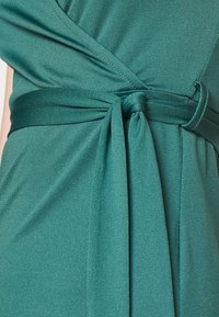 Robe portefeuille turquoise en tissu doux et texturé, avec une large ceinture et un nœud noué sur le côté pour une silhouette ajustée.