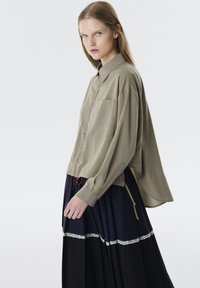 Licht olijfgroene blouse met een kraag, lange mouwen en één zak. Gecombineerd met een gelaagde zwarte en marineblauwe rok met kantafwerking.