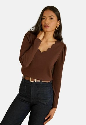 Braunes Langarmshirt mit gewelltem Ausschnitt, kombiniert mit dunklen Jeans. Glatte Stoffstruktur; Modell trägt goldene Creolen.