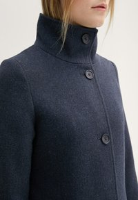 Sötétkék gyapjú kabát magas gallérral, két sötét gombbal, texturált anyaggal és karcsú fazonnal.
