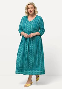 Vestido maxi de color verde azulado con mangas tres cuartos, escote en V y estampados florales. La tela es suave; presenta un cuerpo ajustado y una falda fluida.