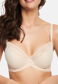 Beige bra with lace detailing, soft cups, and thin adjustable straps. Features a small bow at the center and textured edges.