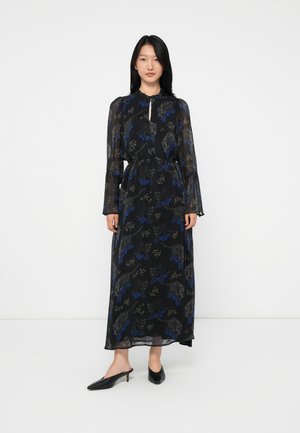 Donna in piedi che indossa un vestito maxi lungo con maniche trasparenti nere, decorato con motivi astratti blu e grigi, e mule nere con tacco.