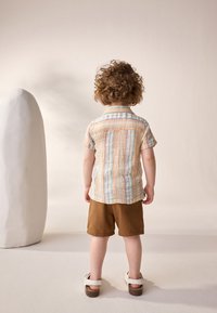 Short-sleeve, multi-striped shirt in pastel colors, brown shorts, and white sandals on a child facing away from a sculptural background.