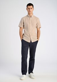 Ljusbeige kortärmad skjorta med raka sidor, kombinerad med marinblå byxor och vita sneakers, stående mot en ljus bakgrund.